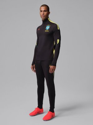 Homme debout portant un survêtement d'entraînement noir du Brésil avec des accents jaunes et des crampons de football rouge vif sur fond gris.