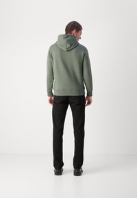 Sudadera verde con un ajuste relajado, que presenta una capucha con cordón y puños de canalé, combinada con pantalones negros y calzado negro.