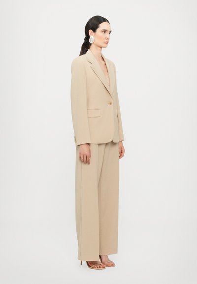 Beige op maat gemaakt pak met een blazer met één knoop en wijde broekspijpen, gemaakt van gladde stof. Vergezeld van beige hakken met bandjes.