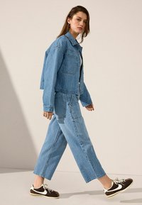 Chaqueta de mezclilla azul claro con rayas verticales, combinada con pantalones vaqueros de corte holgado a rayas y zapatillas marrones con acentos blancos, llevada por un modelo.