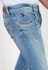 Vaqueros azul claro con acabado desgastado, cinco bolsillos y costuras reforzadas. Presenta un parche de cuero marrón y una pequeña etiqueta con el logo cerca de la cintura.