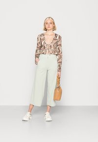 Blusa em padrão espiral castanho e creme com frente amarrada, calças largas verde claro, ténis creme e uma pequena bolsa bege.