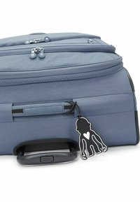 Valise bleue avec une finition texturée, double zip, poignée rétractable et une étiquette en forme de personnage noire avec un détail en cœur.