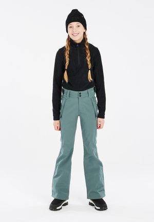Pantalones de esquí verdes con una textura suave, cierre de doble botón y bolsillos con cremallera, combinados con una camiseta de manga larga negra de canalé y un gorro de lana.