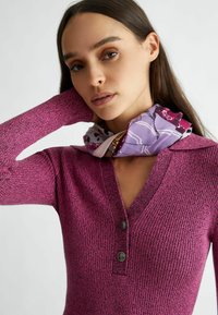 Pullover magenta aderente con texture a costine, dotato di scollatura a V e due bottoni scuri al colletto. La sciarpa abbottonata aggiunge un accento a fantasia.