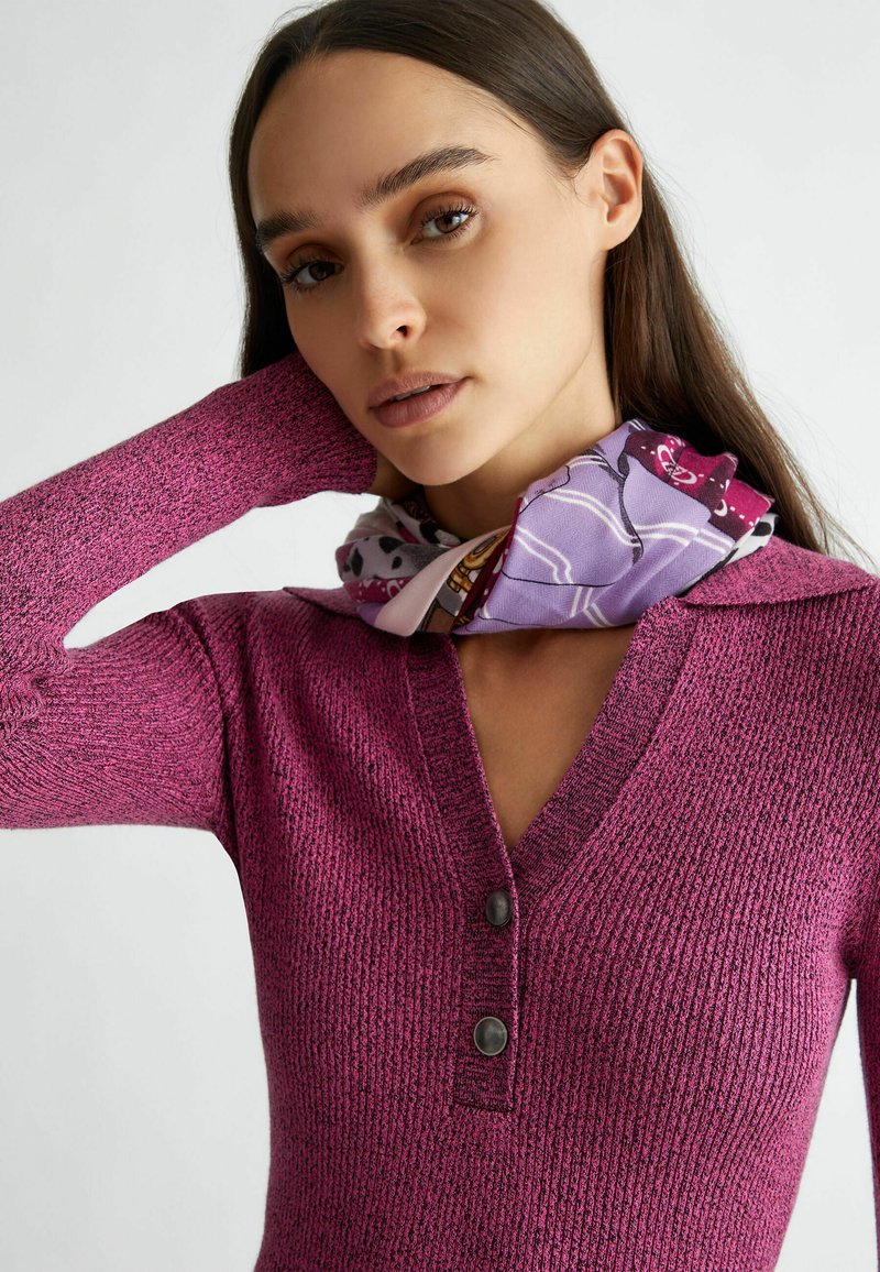 Pullover magenta aderente con texture a costine, dotato di scollatura a V e due bottoni scuri al colletto. La sciarpa abbottonata aggiunge un accento a fantasia.