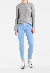 Šedý pletený svetr s ribkovanou texturou, světle modré skinny džíny a lesklé stříbrné kotníkové boty. Jednoduchý design, přiléhavý styl a živé barvy.