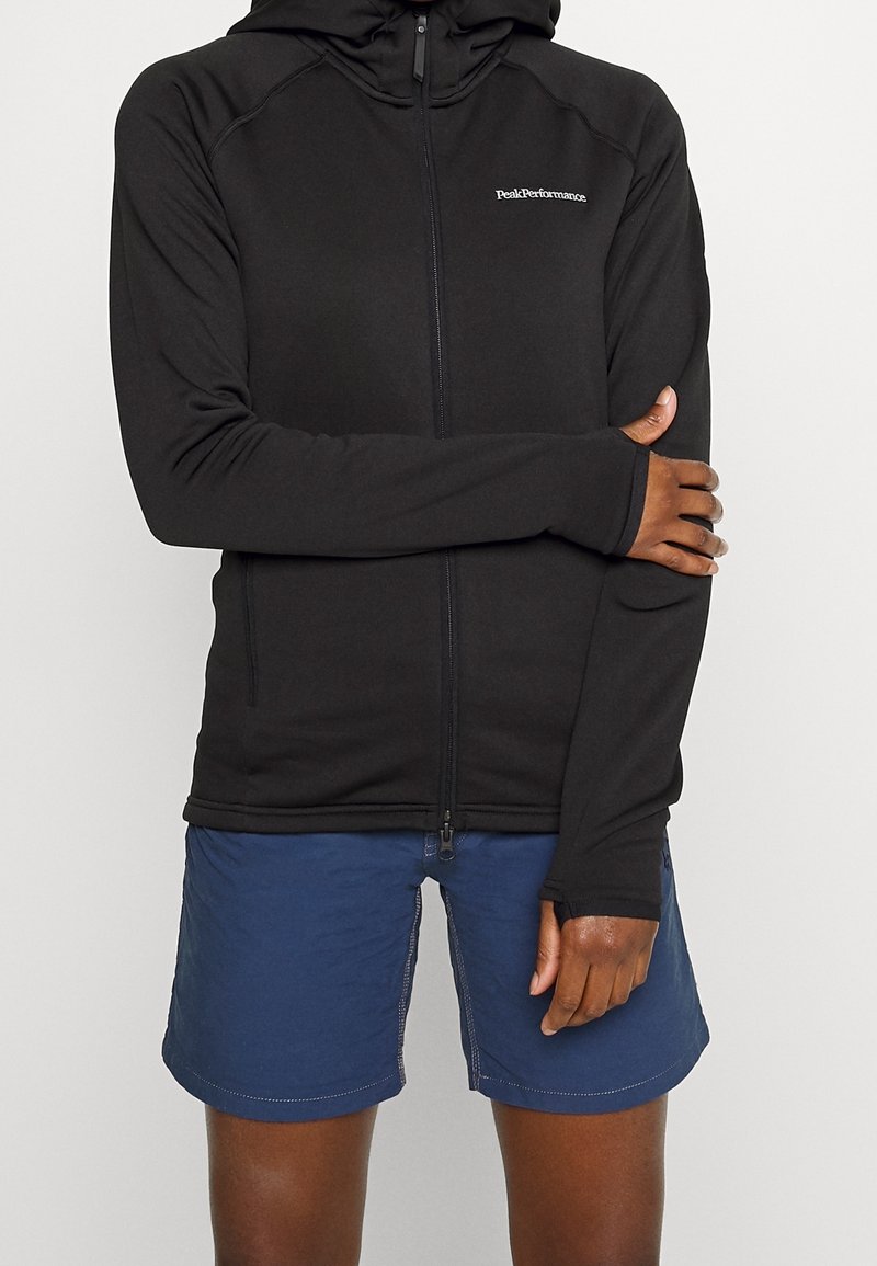 Sudadera negra con cremallera y corte ajustado, que incluye capucha y puños con orificio para el pulgar. Combinada con pantalones cortos azul marino, exhibiendo un estilo casual al aire libre.