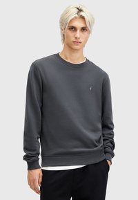 Sudadera de cuello redondo en un suave tejido gris oscuro, con puños y dobladillo acanalados. Pequeño logo bordado en el pecho. Superpuesta a una camiseta blanca.
