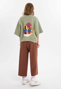 Olivgrön oversized sweatshirt med korta ärmar, med en färgglad grafisk design på baksidan. Matcher med löst sittande bruna byxor och vita sneakers.