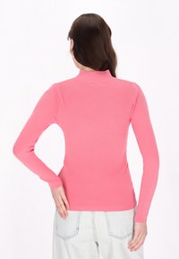 Tricou cu maneci lungi, cu refile, de culoare roz aprins, cu guler înalt. Materialul pare elastic. Asortat cu blugi deschiși la culoare, cu talie înaltă.