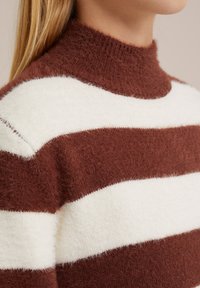 Hnedo-bielo pruhovaný sveter s vysokým, huňatým golierom. Textúra vyzerá mäkko, s širokými horizontálnymi pruhmi a útulným dizajnom.