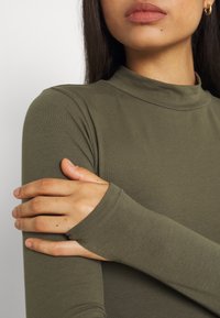 Olivenfarbener Langarm-Oberteil mit hohem Kragen und tailliertem Schnitt. Weicher, dehnbarer Stoff mit verlängerten Bündchen, die die Handgelenke bedecken.