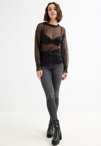 Top negro transparente de manga larga con textura acanalada y lentejuelas, superpuesto sobre un sujetador negro. Combinado con jeans ajustados oscuros y botas negras.