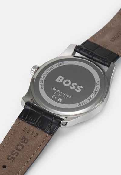 Fond de montre en acier inoxydable avec gravé "BOSS", étiquette résistant à l'eau ; bracelet en cuir noir texturé ; dessous tan avec logo embossé.