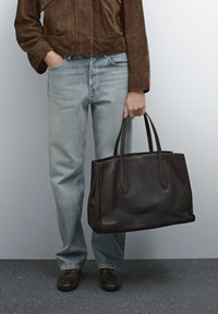 Braune Leder-Shopper-Tasche mit zwei langen Griffen, strukturierter Form, glatter Textur und dezenten Nähten, gehalten von einer Person in hellen Jeans.
