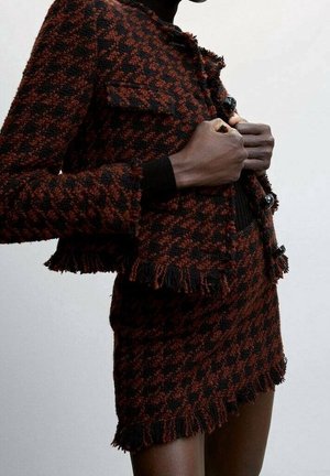 Personne à la peau foncée portant une veste marron et noire à motif pied-de-poule et une jupe assortie avec des détails en franges, en train de boutonner les boutons de la veste.