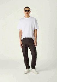 T-shirt branco de manga curta, calças cargo pretas com bolsos laterais e tênis brancos. O modelo usa óculos de sol e está posicionado contra um fundo neutro.