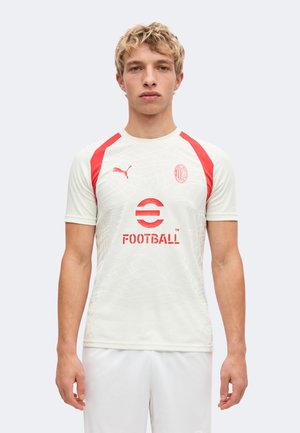 Млад мъж, облечен в бяла и червена футболна фланелка на AC Milan с лого на Puma и текст "Football", стоящ срещу еднообразен светъл фон.