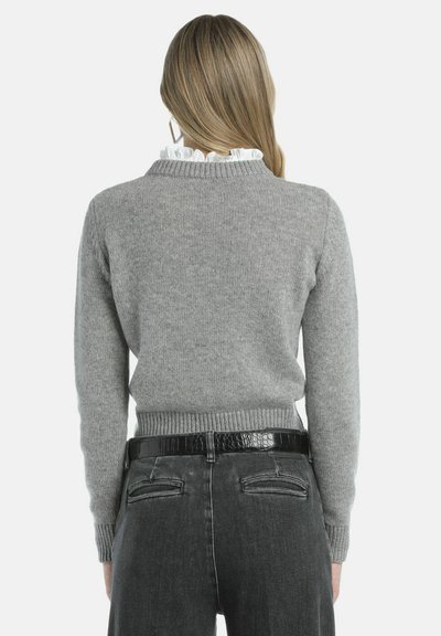 Gemêleerd gebreid vest met een geribde kraag en manchetten, met een witte ruchesdetail bij de hals; gecombineerd met zwarte high-waisted jeans en een glanzende riem.