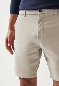 Shorts en coton beige clair avec un devant plat, des poches latérales et une fermeture à bouton. Caractéristiques d'une texture lisse et d'une coupe ajustée.