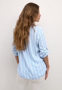 Camisa de botones azul clara con rayas verticales blancas, con mangas arremangadas y dobladillo curvado, fabricada en una tela suave.