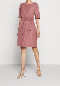 Růžové šaty s krátkým rukávem a detailem vázání v pase, vyrobené z hladké tkaniny, s kulatým výstřihem a dlouhou po kolena.