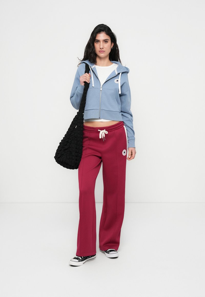 Sweat à capuche bleu raccourci avec cordons blancs, pantalon de survêtement bordeaux avec accents blancs, et sac à épaule noir texturé. Des baskets noires complètent le look.