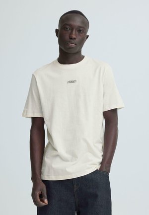 Homme debout, une main dans la poche, portant un t-shirt beige clair avec un petit logo et un jean bleu foncé, sur un fond clair uni.