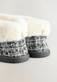 Black and white tweed slippers with a fluffy white faux fur cuff, rounded shape, and black rubber sole, featuring a small accent tag.