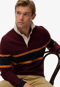 Mørkerød langærmet polo shirt med cremefarvet krave; har vandrette sorte og orange striber samt broderet logo på brystet.