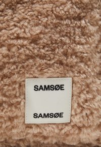 Tissu beige flou avec une surface texturée et pelucheuse. Une étiquette blanche sur laquelle est imprimé "SAMØSØ" en noir est visible.