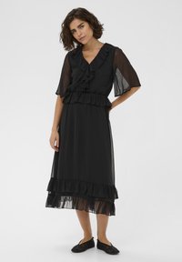 Robe noire en tissu léger avec des détails froncés le long de l'encolure, des manches et de l'ourlet. Silhouette évasée fluide avec des accents transparents.