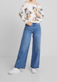 Top floreale bianco senza spalle con maniche lunghe; jeans blu in denim a vita alta e gamba larga; sneakers bianche completano l'outfit.
