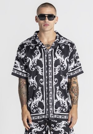 Muž s krátkymi vlasmi a slnečnými okuliarmi nosí čierno-bielu krátkosleevé vzorované tričko a zladené šortky, zobrazujúc tetovania na rukách a hrudi.