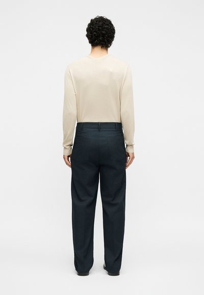 Άτομο με σγουρά σκούρα μαλλιά, ντυμένο με μπεζ μακρυμάνικη μπλούζα και φαρδιά μαύρα παντελόνια, στέκεται αντικρίζοντας ένα απλό λευκό φόντο.