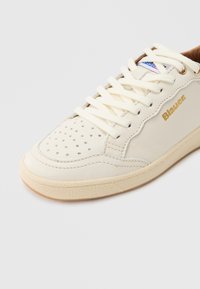 Pantof sport din piele crem cu vârf perforat, șireturi albe, găuri pentru șireturi aurii și logo-ul "Blauer" auriu pe panoul lateral.