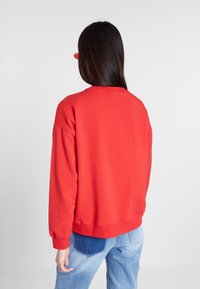 Rode sweatshirt met een ronde halslijn, ribgebreide manchetten en onderrand. Zacht materiaal. G gedragen met blauwe jeans. De achtergrond is effen wit.