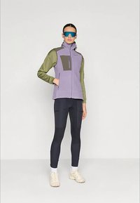Γιλέκο από φλις σε μπλοκ χρωμάτων λιλά και ελιάς, με τσέπη στο στήθος με φερμουάρ και ψηλό γιακά. Το μοντέλο φορά σκουρόχρωμο κολάν και μπεζ αθλητικά παπούτσια.