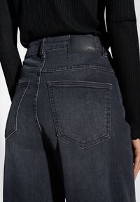 Jean en denim foncé taille haute avec une texture lisse, comportant deux poches plaquées au dos et une étiquette en cuir à la taille.