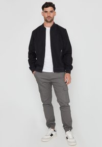 Schwarze, leichte Bomberjacke über einem weißen T-Shirt, kombiniert mit grauen Cargo-Hosen mit Seitentaschen und weißen Sneakers mit schwarzen Akzenten.