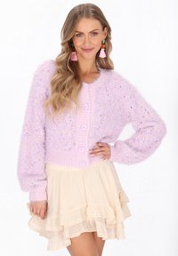 Kobieta uśmiechająca się, ubrana w fioletowy, futrzany sweter na guziki i kremową spódnicę z falbanami, z długimi, falującymi włosami i różowymi frędzlami w uszach.