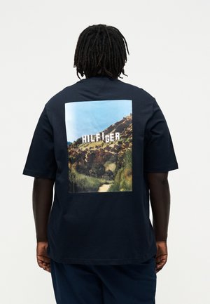 Person med mørk hud og dreadlocks bærer en mørk marineblå t-shirt med en bakke og ordet "HILFIGER" i store hvide bogstaver.