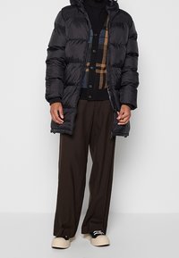 Personne portant une doudoune noire sur un cardigan à motifs, un pantalon large marron et des baskets beiges avec des détails noirs.