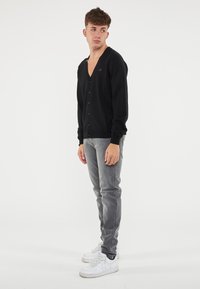 Cardigan nero con scollo a V, bottoni frontali e maniche lunghe, abbinato a jeans grigi e sneakers bianche, su uno sfondo neutro.