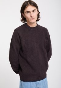 Jeune homme aux cheveux bruns mi-longs, portant un pull en maille marron foncé et un jean bleu clair sur un fond clair uni.