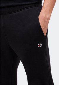 Main dans la poche portant un pantalon noir avec le logo Champion sur la cuisse sur un fond clair uni.