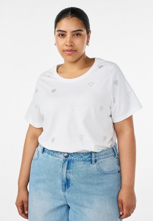 Woman wearing a white short-sleeve shirt with small heart patterns and light blue high-waisted jeans, standing against a plain background.