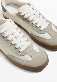 Beige Wildleder- und Leder-Sneaker mit weißen Schnürsenkeln, ausgestattet mit einer strukturierten Gummisohle und sportlichen Designelementen, darunter kontrastierende Einsätze.