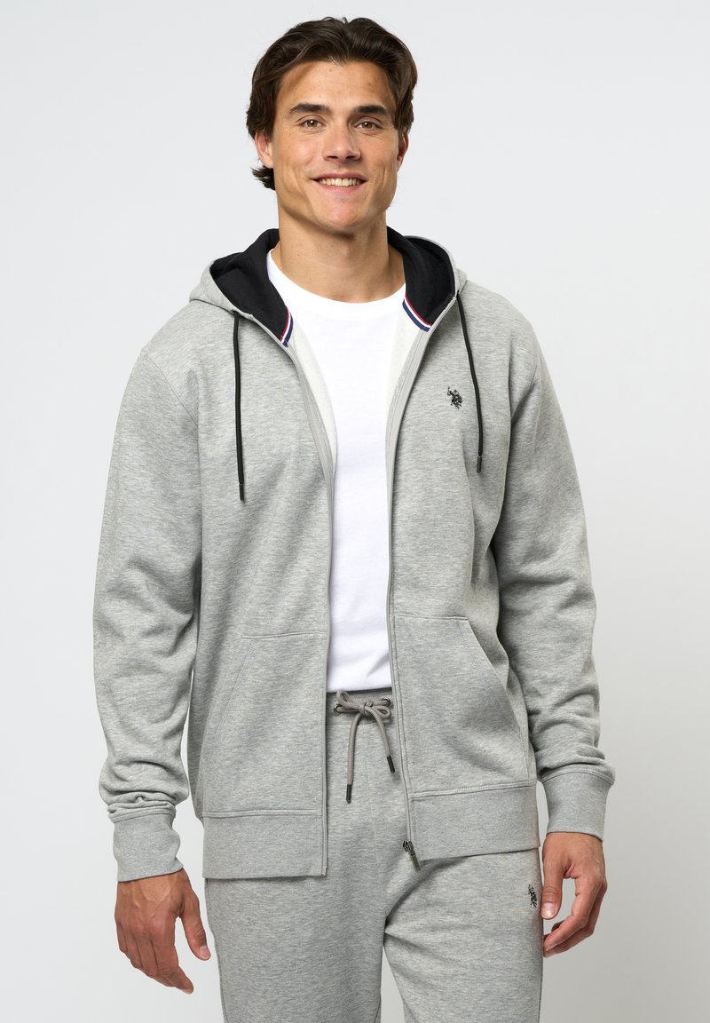 Sivi zip-up dukserica s crnim podstavom, s kengurskom džepom, vezicama i malim logotipom na prsima. Uparena s odgovarajućim hlačama.